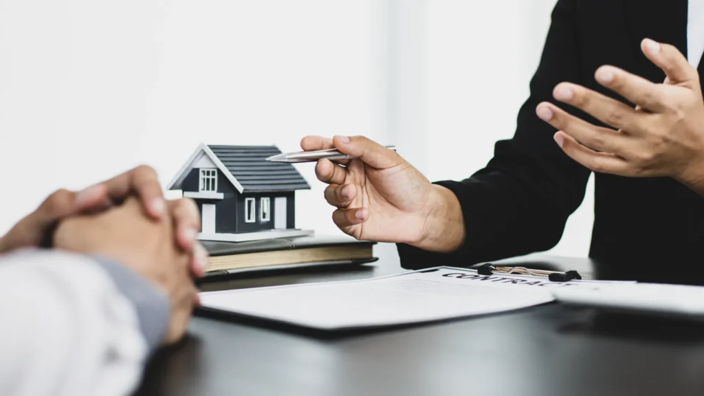 A close-up of two people discussing a contract with a miniature house model on the table, representing estate planning.